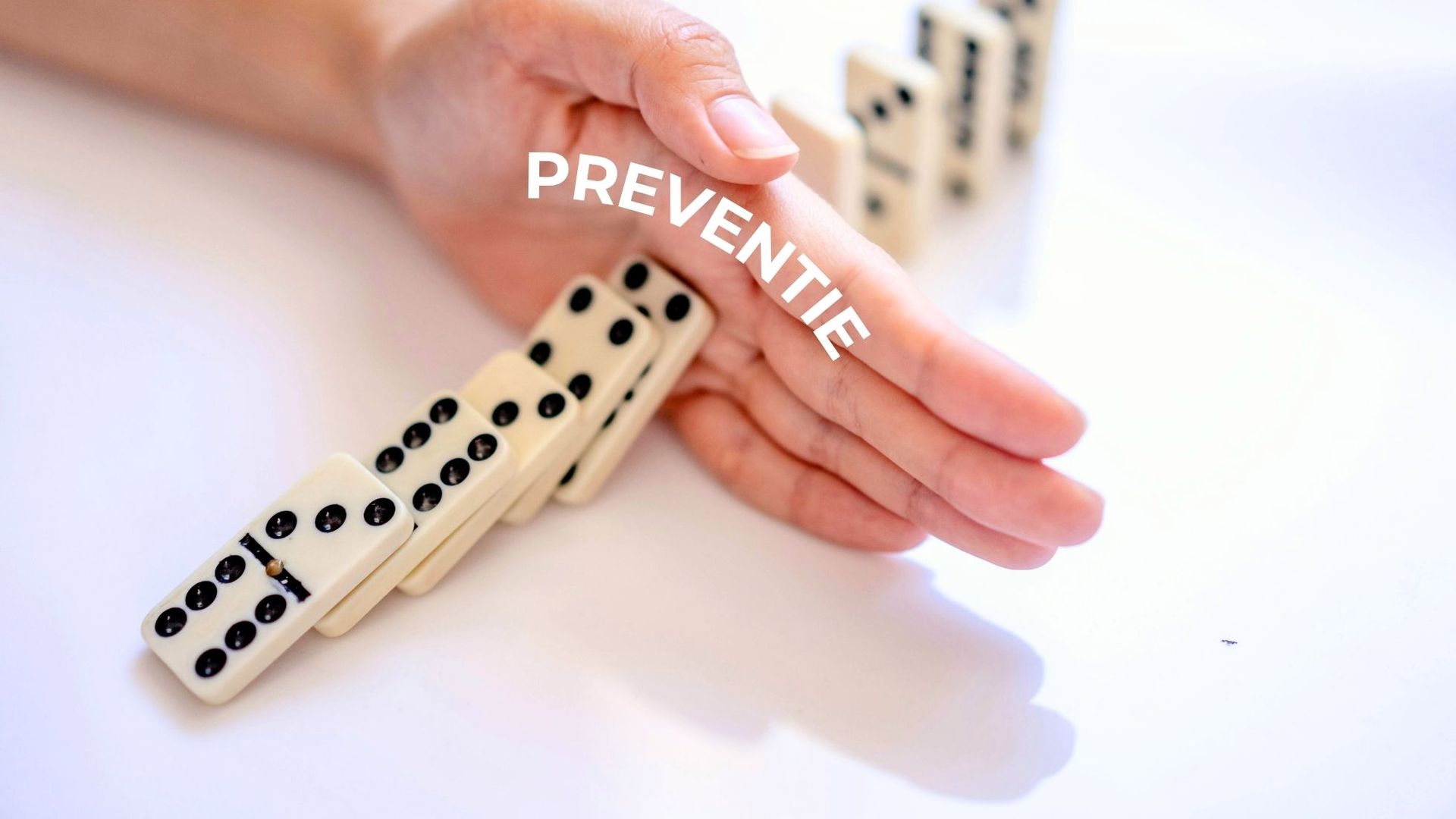 Een rij dominostenen is omgevallen tegen iemands hand waar burn-out preventie op staat met daarachter nog staande domino's.