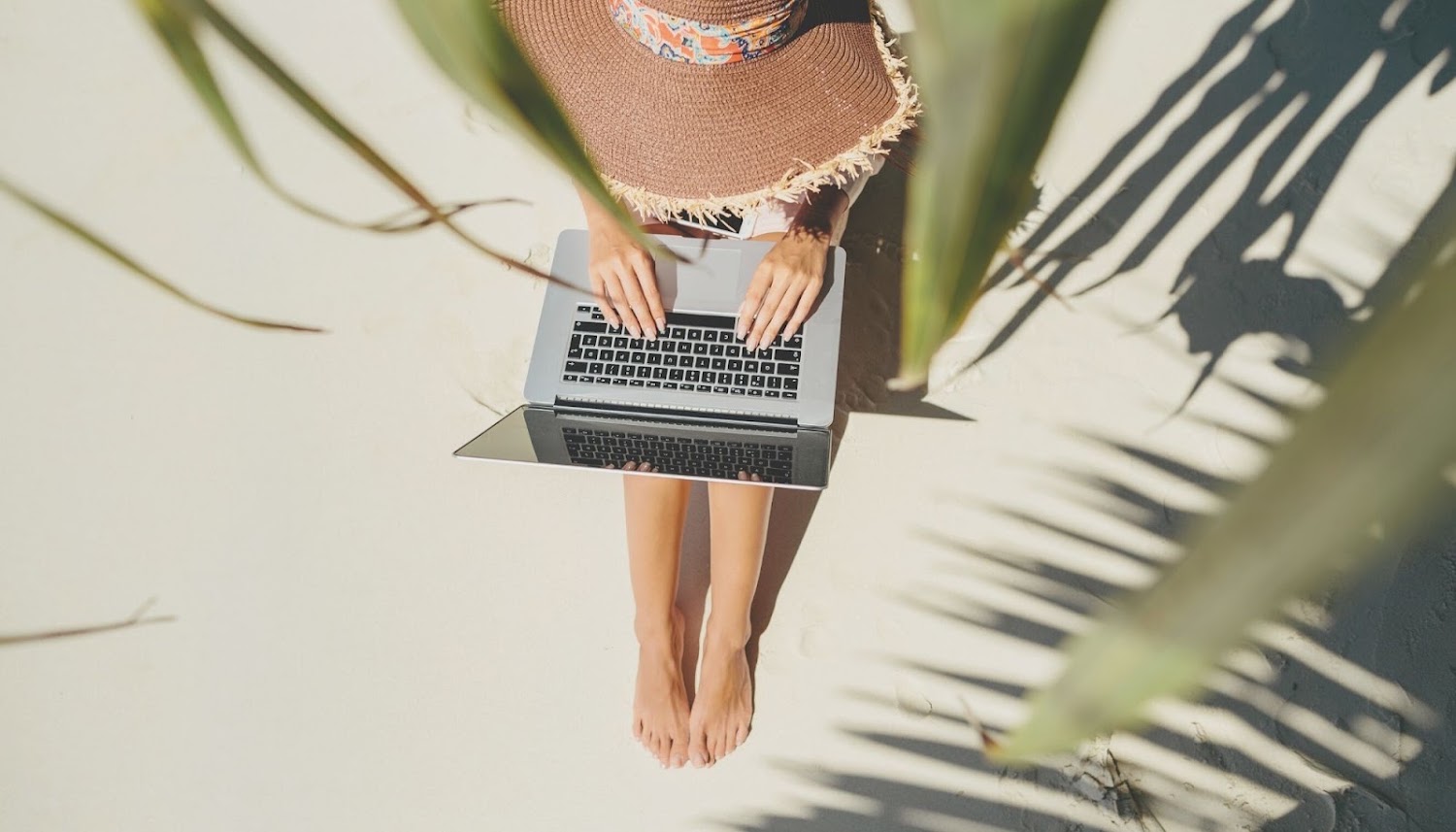 Iemand met een hoed zit op het strand te werken achter haar laptop, zittend op het strand want dat is een voordeel van het ondernemerschap!